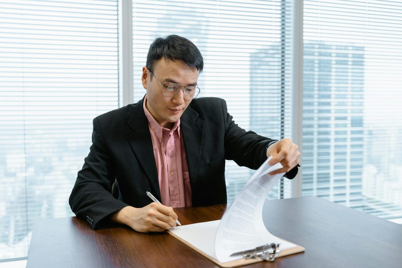 Asian businessman in formal wear signs paperwork at office desk.
